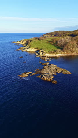 A rocky shoreline extends into the water with a green hill nearby. The sea is calm, and a clear sky is visible. This scene is captured during the day.
