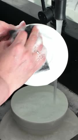 Hands are washing dishes in a sink, rinsing a bowl and a mug under running water, followed by scrubbing with a sponge, showcasing the cleaning process in action