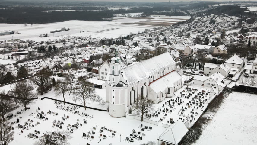 Leftward orbiting drone around snowy Elchingen church with cemetery, village homes, snowy fields under overcast skies. Perfect for travel docs, ads, reveals.