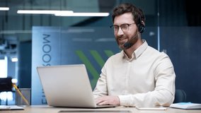 Male call center operator wearing headset working on laptop in modern office. Corporate customer support agent talking to client on helpline, providing service assistance, telesales, technical support - Powered by Shutterstock - Get 15% off with code: PIKWIZARD15