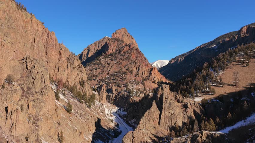 A cinematic aerial vista highlights a sharp mountain peak rising above steep rocky canyon walls. Dramatic deep shadows and winter snow showcase the immense scale of this rugged mountain environment.