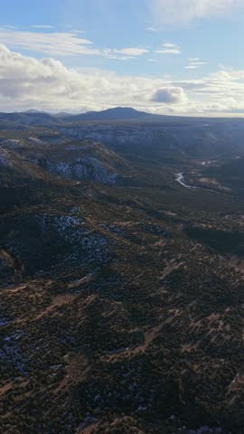 A cinematic vertical aerial view captures a river winding through a vast Southwest canyon. Light snow dusts the rugged high desert hills under a bright sky, ideal for mobile storytelling.