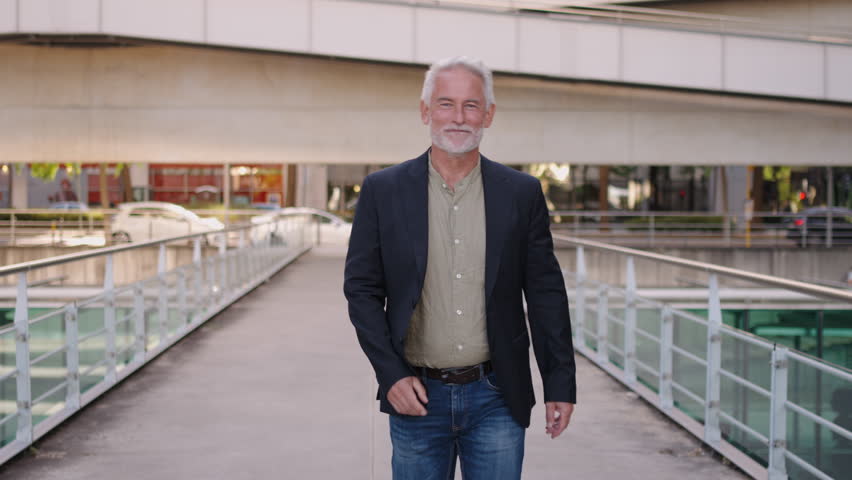 Confident senior businessman walking on urban footbridge in daylight near modern office buildings, expressing success, positivity and stylish professional attitude