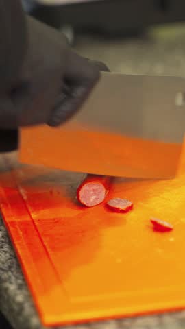 Close-up view of grilled sausage slices being expertly cut on an orange cutting board. The focus is on texture and color, highlighting the artistry of cooking.