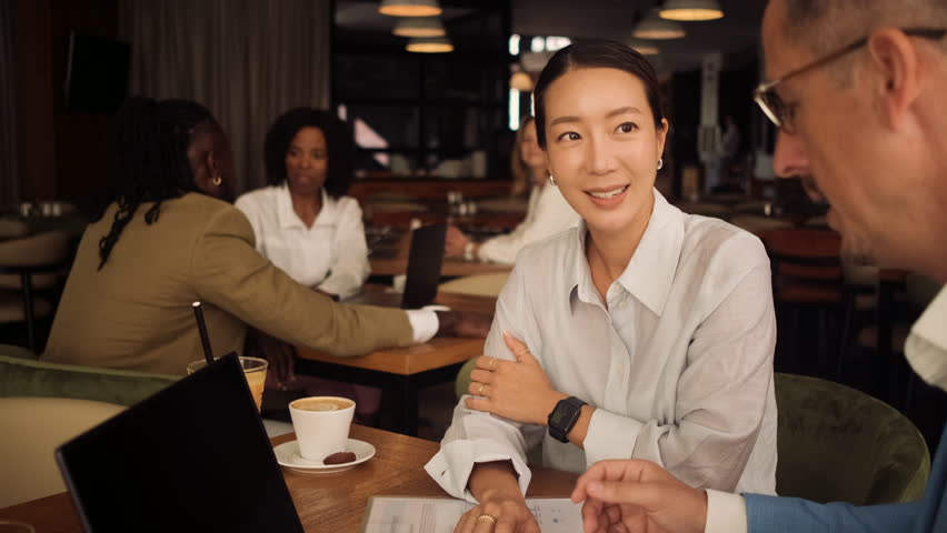 Asian woman and coworker talking about work while using laptop in modern coffee shop.