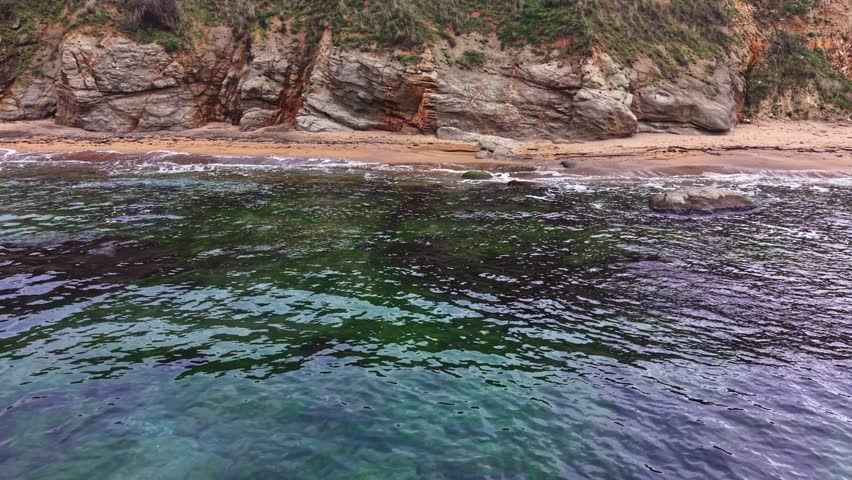 This aerial shot captures a rocky coastline meeting calm waters. The beach is visible at low tide, showcasing sand and gentle waves lapping at the shore.