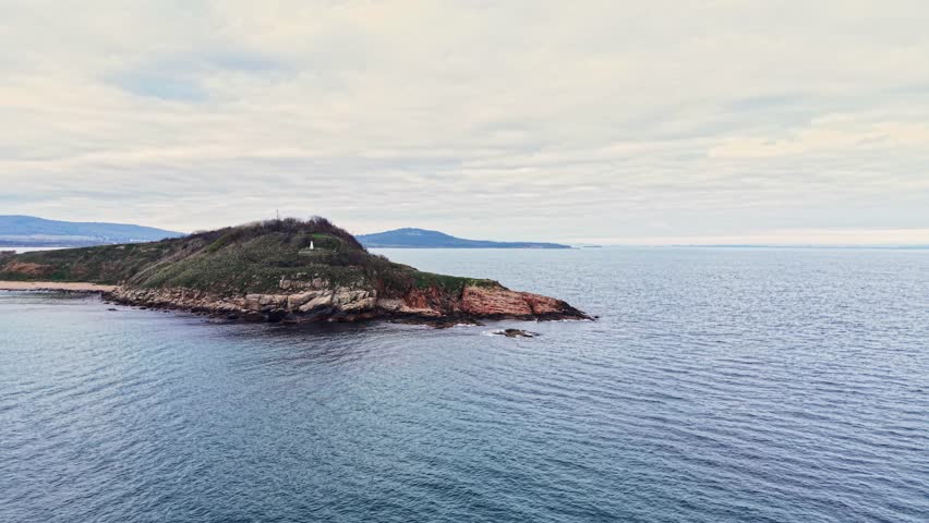 An aerial perspective captures a rocky island surrounded by tranquil ocean waters, showcasing the lush greenery and distant hills along the serene coastline.
