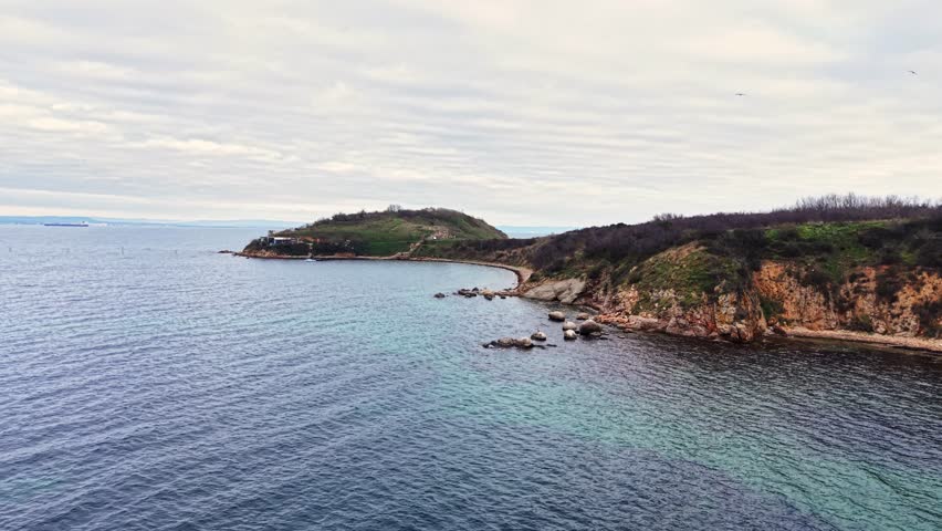 An aerial view captures the rocky coastline, green hills, and calm blue waters. The scene is peaceful, showcasing nature's beauty during a cloudy sunset.