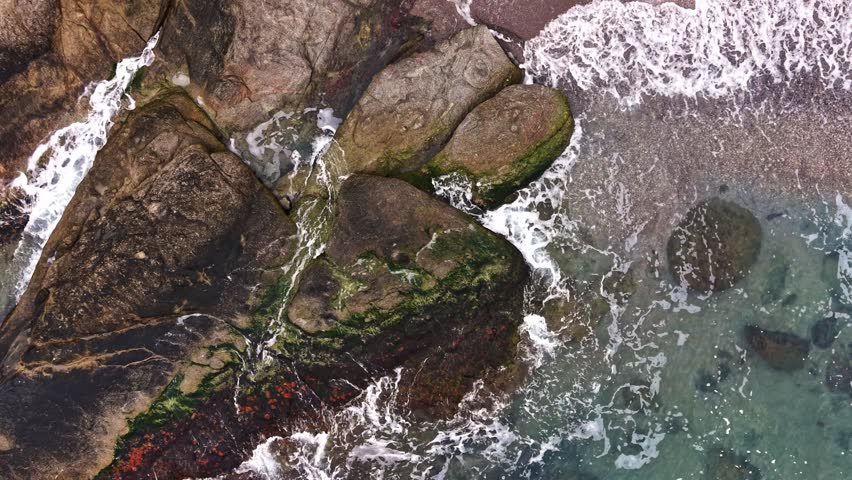 Aerial view showcasing a rugged coastal shoreline. Waves softly crash against rocky formations, revealing clear water and patches of green moss.