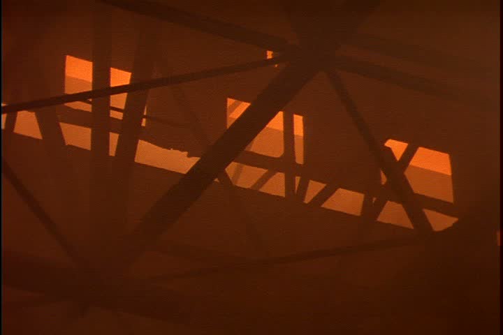 Steel works in Nizhny-Tagil, Russia. WS rafters and windows obscured by thick orange smoke, camera tilts down to molten iron being poured from ladle into open hearth furnace.