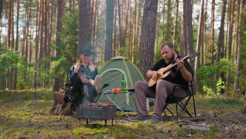 Young couple relaxing together camping in the woods, man playing acoustic guitar and woman listening and grilling steak on barbecue this weekend