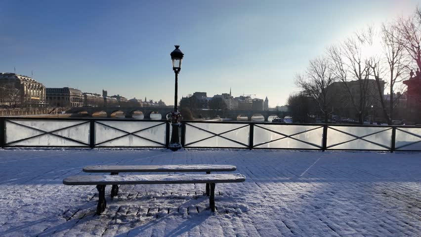 A snow-covered bench and bridge railing frame the Seine river from Pont des Arts in Paris on a bright winter morning.