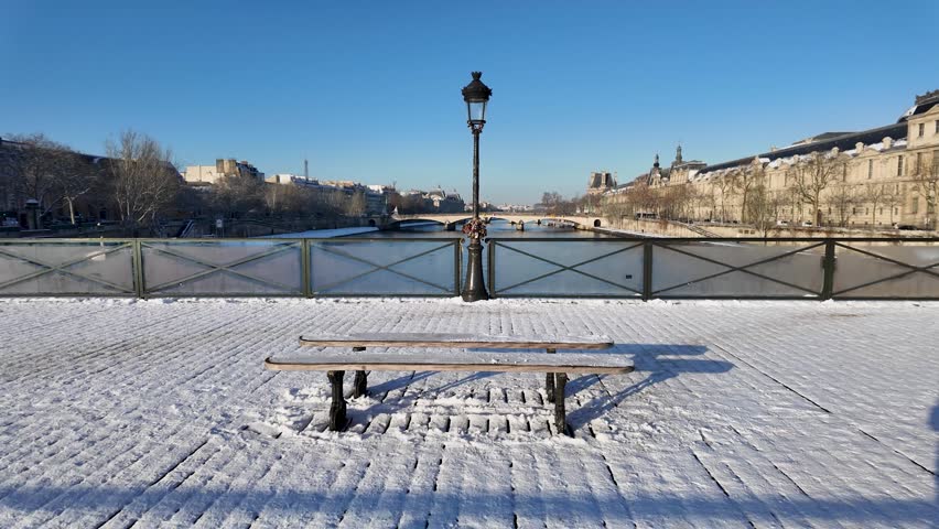 A snow-covered bench and bridge railing frame the Seine river from Pont des Arts in Paris on a bright winter morning.