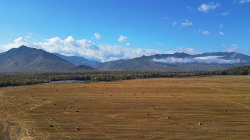 High-altitude drone shot over a vast autumn field with scattered hay bales, with a wide mountain range on the horizon under a blue sky with clouds. Altai Republic, Russia.