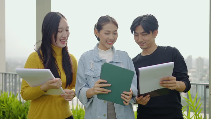 Group of diverse young Asian business colleagues brainstorming project on urban rooftop balcony using tablet and documents. Professionals collaborating outdoors with a city skyline background.