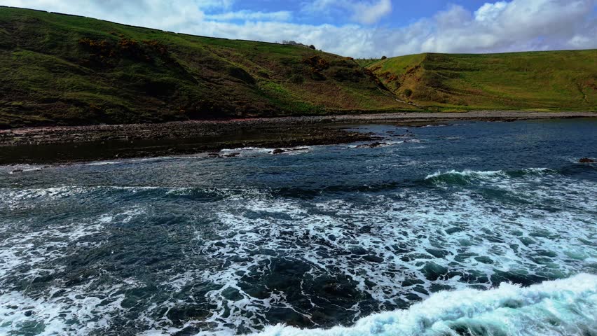Waves crashing onto rocky shoreline on North Sea coast in Scotland. Swells foaming across shallows near grassy hills. Tides surging toward pebbled beach under cloudy sky. Currents pushing through