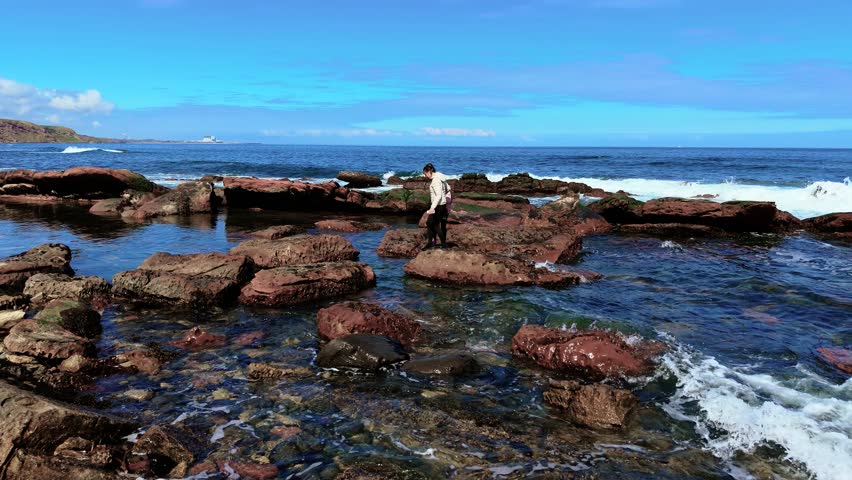 Mother and daughter jumping across rocks on sea shore. Family leaping over stones by ocean during coastal walk. Woman and girl playing near waves and skipping on seaside boulders