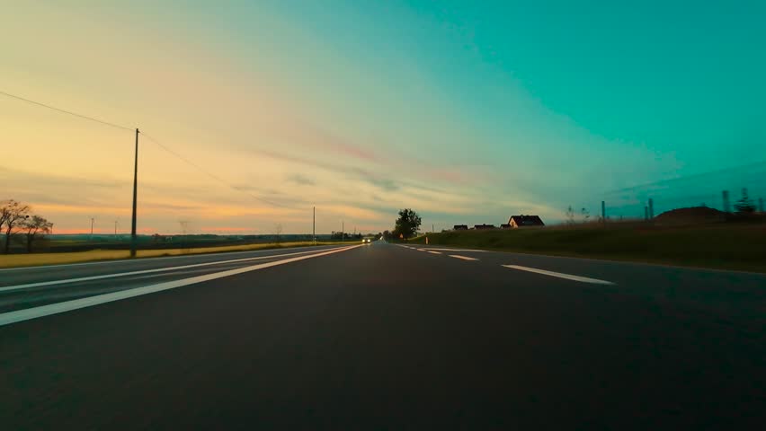 POV driving on asphalt road at sunset with headlights on. Point of view moving along paved highway during dusk with lights shining. Sunset journey cruising down smooth roadway with beams illuminating