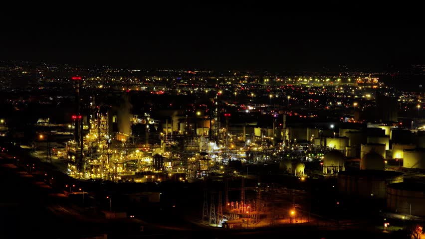 Thessaloniki, Greece — Drone footage and photographs capture the illuminated industrial complex of Hellenic Petroleum at night, showcasing active refinery units, storage tanks, chimneys, and the surrounding urban and port infrastructure.