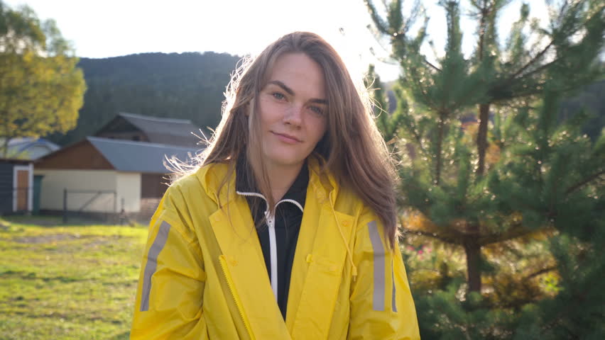 Portrait of a young caucasian woman with freckles wearing a yellow raincoat. She is standing outdoors with sun flare behind her