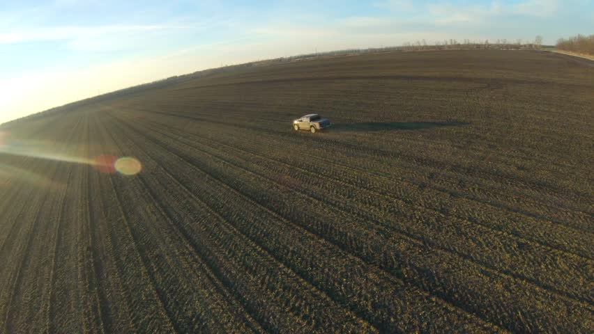 Off road vehicle riding along ploughed meadow. Pickup truck driving through plowed field at sunset time. Black car going on route with sunlight at background. Concept of agronomy farming. Aerial shot