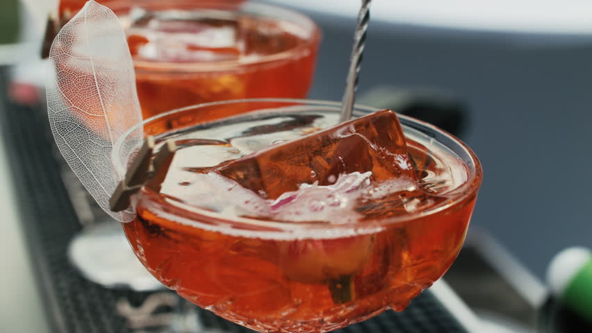 Orange cocktail with ice cube and metal stirrer in coupe glass
