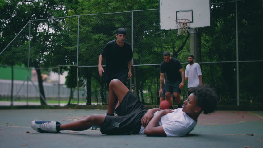 Hispanic and African descent men on outdoor basketball court, one helping the other up after falling during tense game, gesture of reconciliation and sportsmanship