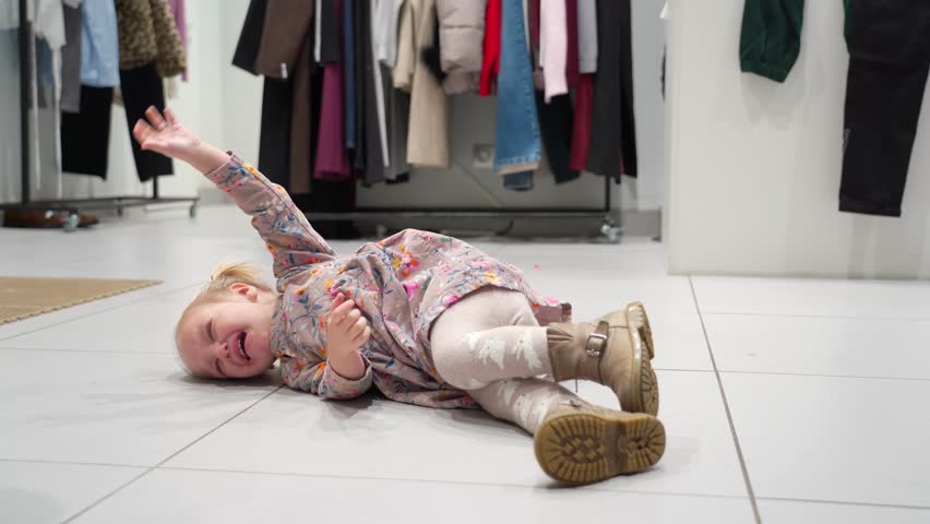 Young child in store throws tantrum on floor, upset and crying while lying down amidst clothing racks