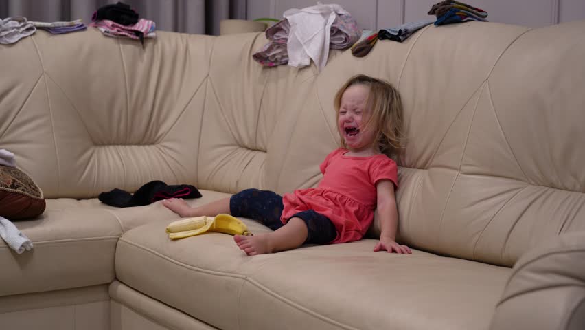 Upset blonde toddler girl sitting on messy beige sofa throwing fit and crying hysterically during challenging childhood moment