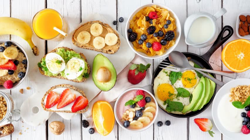 Healthy breakfast table scene with fruit, yogurts, oatmeal, smoothie, nutritious toasts and egg skillet. Slow rotating zoom motion. Overhead view on a white wood background.