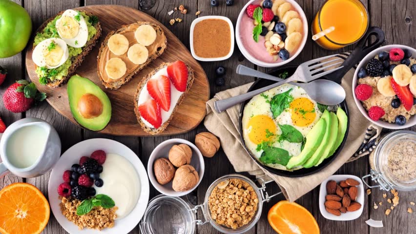 Healthy breakfast table scene with fruit, yogurts, oatmeal, smoothie, nutritious toasts and egg skillet. Slow rotating zoom motion. Top view over a dark wood background.