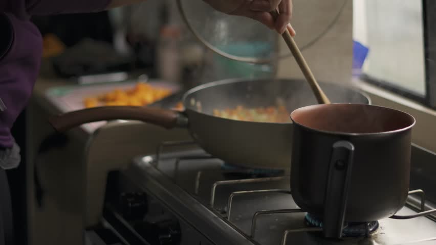 Close-up of a person sautéing chopped vegetables in a large pan on a gas stove inside a motorhome