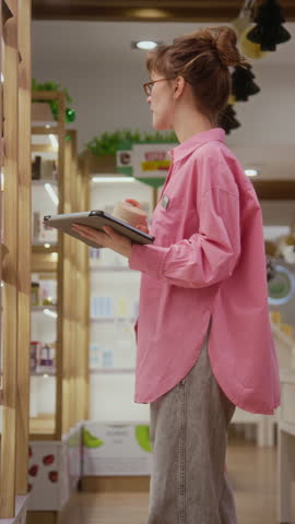 Vertical side view shot of female store worker wearing name badge inspecting skincare stock on shelves using tablet computer at cosmetics store