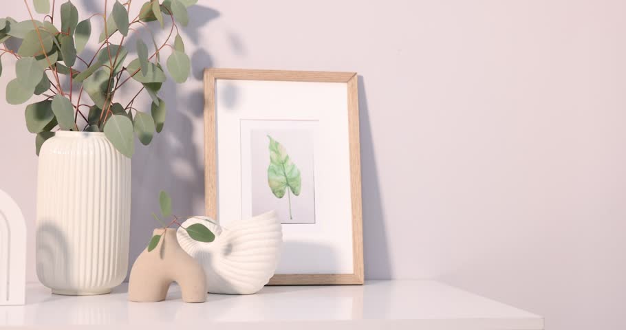 Eucalyptus branches in vases, picture and burning candle on white chest of drawers near light wall, camera moving left and backwards