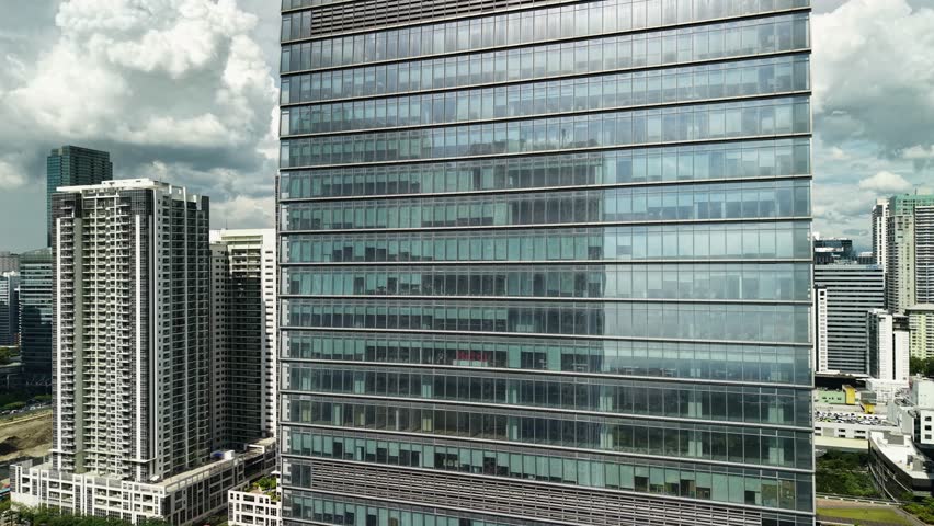 Cinematic aerial footage captures a slow vertical ascent along the sleek glass exterior of a contemporary high-rise office tower in Bonifacio Global City, Manila, Philippines. The camera glides upward near the reflective curtain wall, revealing horizontal metallic louvers that create distinctive bands across the transparent facade. Floor after floor of glass panels pass by, each reflecting the dramatic cloudscape above with white cumulus formations mirrored on the surface. The architectural detail showcases precision engineering with clean lines and geometric patterns formed by window frames and ventilation strips. Surrounding residential and commercial towers populate the background, creating depth in the urban landscape of this premier business district. Natural daylight illuminates the scene while cloud shadows dance across the reflective exterior, adding dynamic visual interest to the composition. The tight framing emphasizes the scale and modern design of the structure, with subtle details of interior offices visible through semi-transparent windows. The atmospheric sky provides a moody backdrop, contrasting with the crisp, contemporary lines of the architecture.