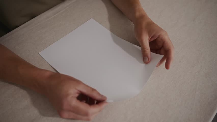 Medium top-down shot showing an adult man carefully folding a paper airplane on a table. Camera captures calm movements and step-by-step paper folding. High quality 4k footage