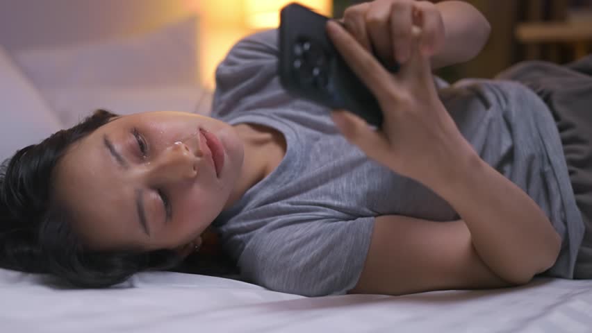 Depressed young Asian woman looking at mobile phone device at night. Frustrated girl lying down on bed in bedroom, feeling heartbroken after breakup, overwhelmed by life problems and financial stress.
