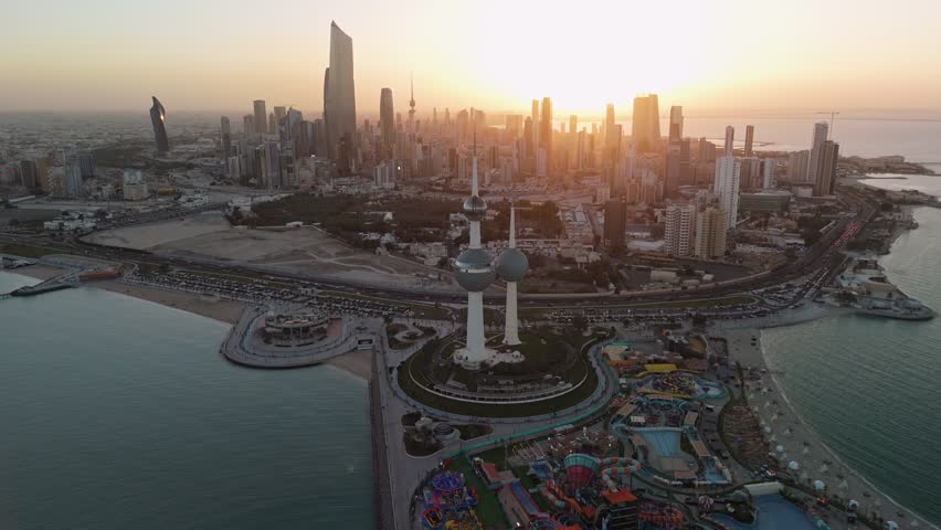 Drone aerial golden hour view of Kuwait Towers with Kuwait City skyline and Al Hamra Tower, coastal highway, seaside park and Arabian Gulf waterfront at sunset.