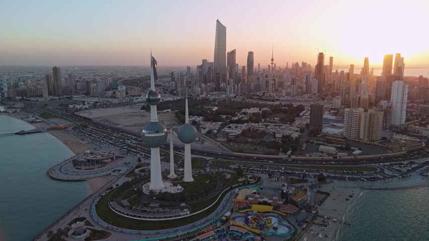 Drone aerial at golden hour of Kuwait Towers and Kuwait City skyline, coastal highway and Arabian Gulf waterfront with seaside park, beach curve, and city traffic.