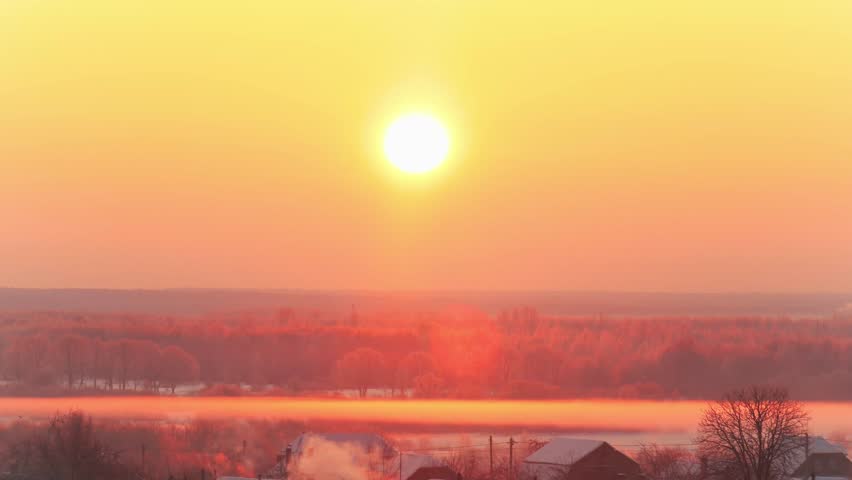 Winter morning landscape with expansive golden sunrise across horizon, sun casting warm glow over frosted trees and rural village. Mist rises from ground, smoke plumes emerge from houses.