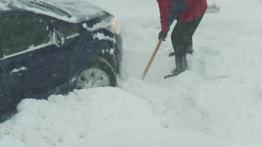 Detailed view of person using shovel to remove extensive snow accumulation from around front wheel of dark blue car. Scene depicts challenging winter weather including active snowfall