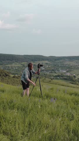 A man sets up a camera on a tripod to take photos in the grassy hill area at sunset.
