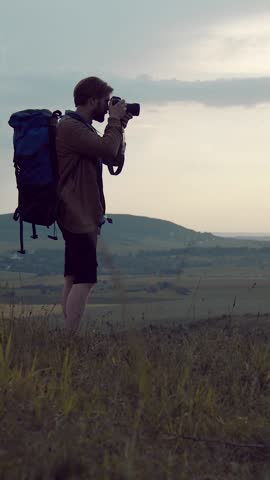 Man stands on hill with camera, photographing landscape during twilight.