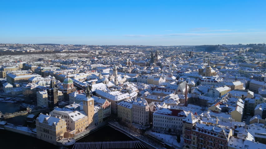 Aerial view of Prague Old Town Square in winter. Snow-covered rooftops, Týn Church, and sunny blue sky. Stunning historic Czech Republic cityscape.