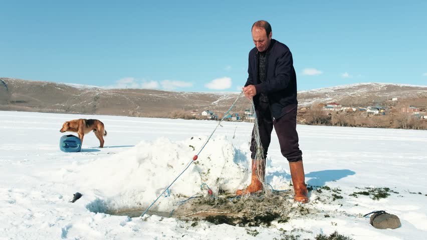 Fisherman setting a fishing net into an ice hole on a frozen lake in Kars, Turkey. Cinematic winter scene showing traditional ice fishing, rural life, cold weather and outdoor lifestyle concept.