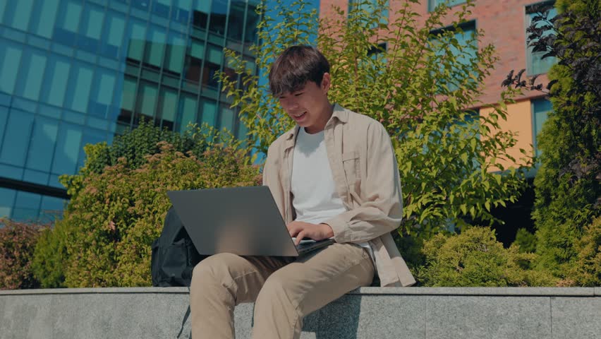 An aspiring student, a young man of Asian Korean descent, engaged in scholarly pursuits, seated on a park bench, intently studying on his laptop in the midst of nature