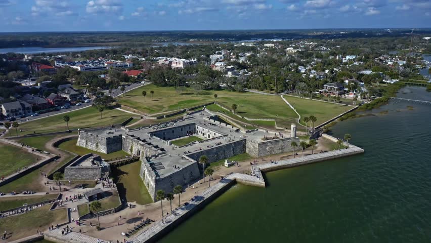 Historic Castillo de San Marcos stands as a large stone fortification in St. Augustine, Florida, surrounded by water, green parkland, and urban buildings on a partly cloudy day.