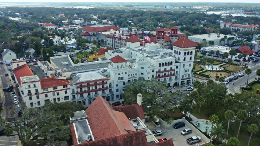 Flagler College in St. Augustine, Florida, features historic Spanish Renaissance architecture with red tiled roofs and a prominent American flag flying, surrounded by lush greenery and urban streets.
