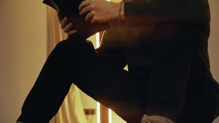 Young man reading the holy bible and praying in a modern church with a neon cross in the background