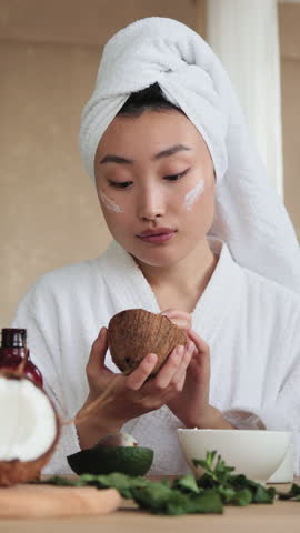 A young woman with a towel on her head and wearing a bathrobe applies cream to her face while holding a coconut